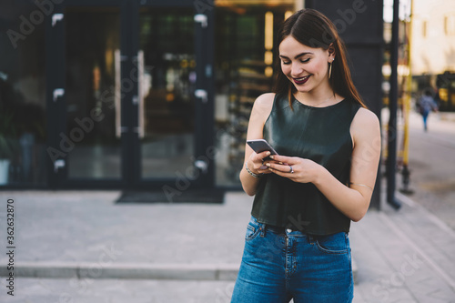 Beautiful smiling hipster girl dressed in fashionable brand clothes having fun while watching interesting video in website on cellular.Young happy student booking online and walking at urban setting