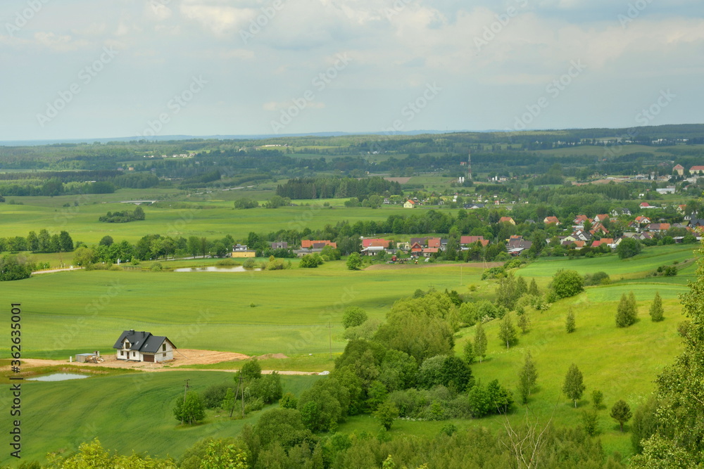 Obraz premium View of a hilly area full or meadows, forests, and small villages seen from the top of a tall hill on a cloudy yet warm summer day on a Polish countryside