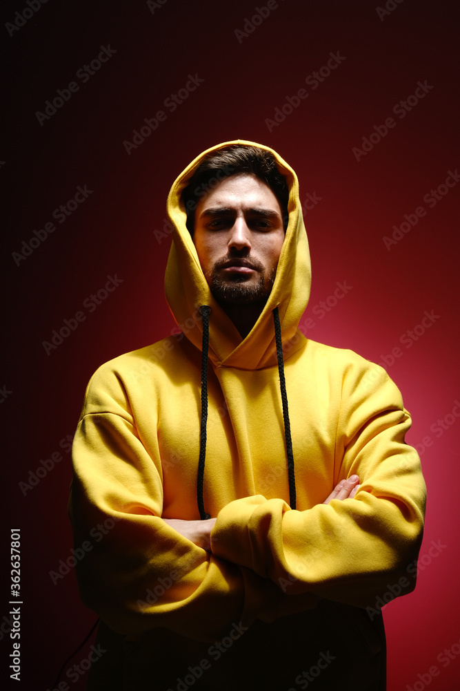 A young man of 25-30 years old in a yellow sweatshirt is standing against a red wall. 