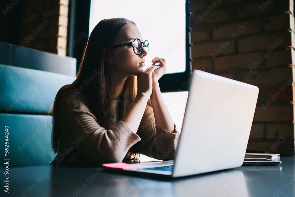 Thoughtful beautiful teenager with trendy eyeglasses with red hair looking out of window and thinking.Pensive redhead woman in spectacles sitting at laptop, computer and pondering on working plans Thoughtful beautiful teenager with trendy eyeglasses with red hair looking out of window and thinking.Pensive redhead woman in spectacles sitting at laptop, computer and pondering on working plans