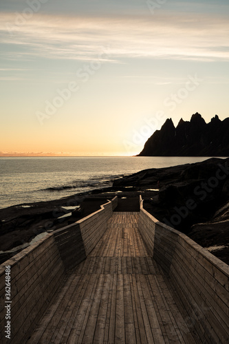 Amazing view during polar day.  Sunset, sharp mountain, calm sea. Mountains background. Wooden path on the rocky coast. Beautiful romantic sky.  "Devil teeth".