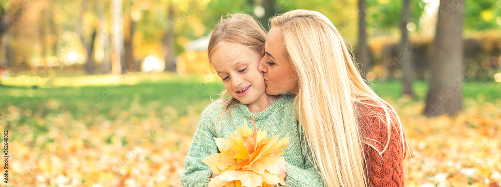 Happy young caucasian mother and little daughter holding autumn yellow leaves sitting and kissing at the park - obrazy, fototapety, plakaty 