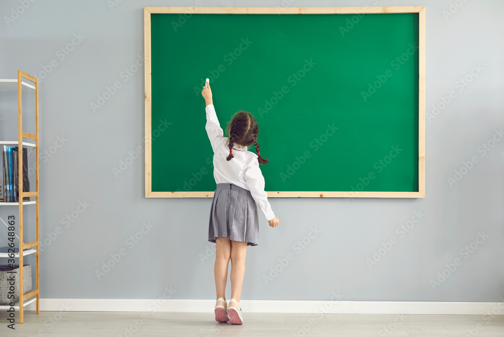 Back view of little girl writing on blackboard with chalk at classroom. Smart first grader answering on lesson at school - obrazy, fototapety, plakaty 