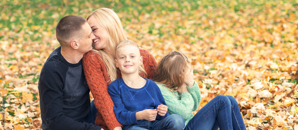 Fototapeta premium Portrait of young family sitting in autumn leaves. Parents kissing and sitting with children in the autumn park