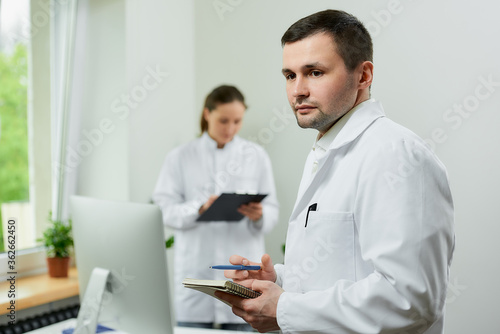 A caucasian doctor is staying with a notebook and a pen in a hospital. A female therapist is doing notes in a clipboard near an all in one computer in a clinic.