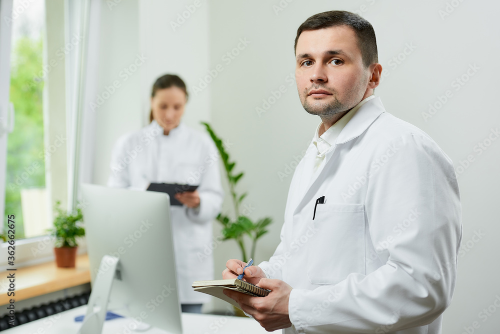Fototapeta premium A serious doctor with a notebook and a pen is thinking about the patient’s medical history in a hospital. A female therapist is doing notes in a clipboard near an all in one computer in a clinic.