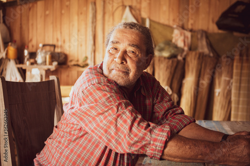 Old artisan in a local community of Garopaba, Brazil. Craftsman resting at his wooden workshop. Elder wearing red plaid shirt