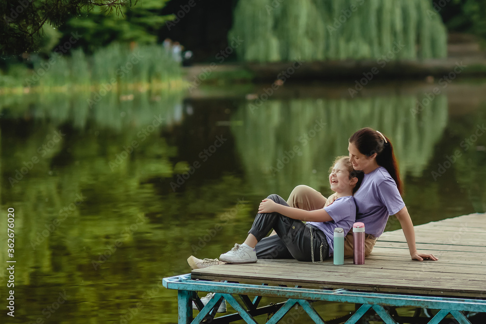 Happy mother and daughter sit on a bridge in the park against the backdrop of a beautiful lake.