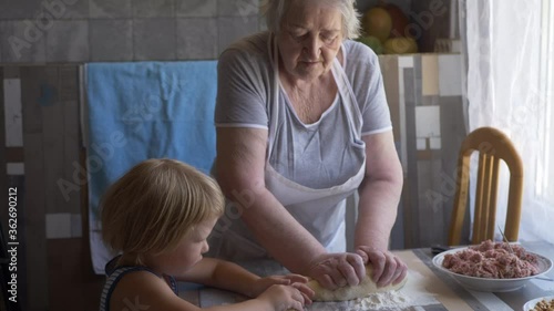 Funny blond boy helps beloved  grandmother in kitchen. elderly woman rolls pastry for pasties and puts minced meat. teenager touches dough. Kid and granny are happy together. shallow depth of field 