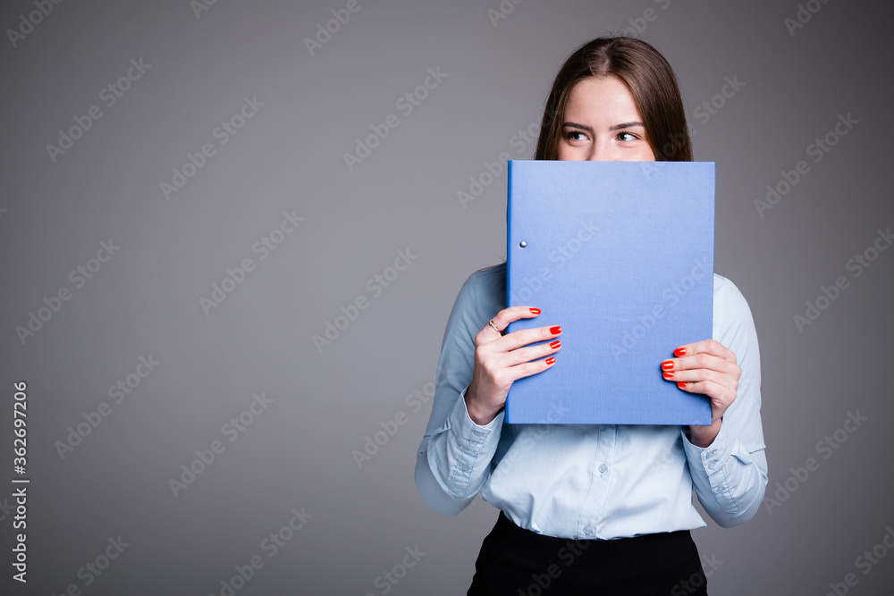 Business portrait of an office worker who is standing on a gray background and peeking out from behind a folder with documents in front of her Business portrait of an office worker who is standing on a gray background and peeking out from behind a folder with documents in front of her