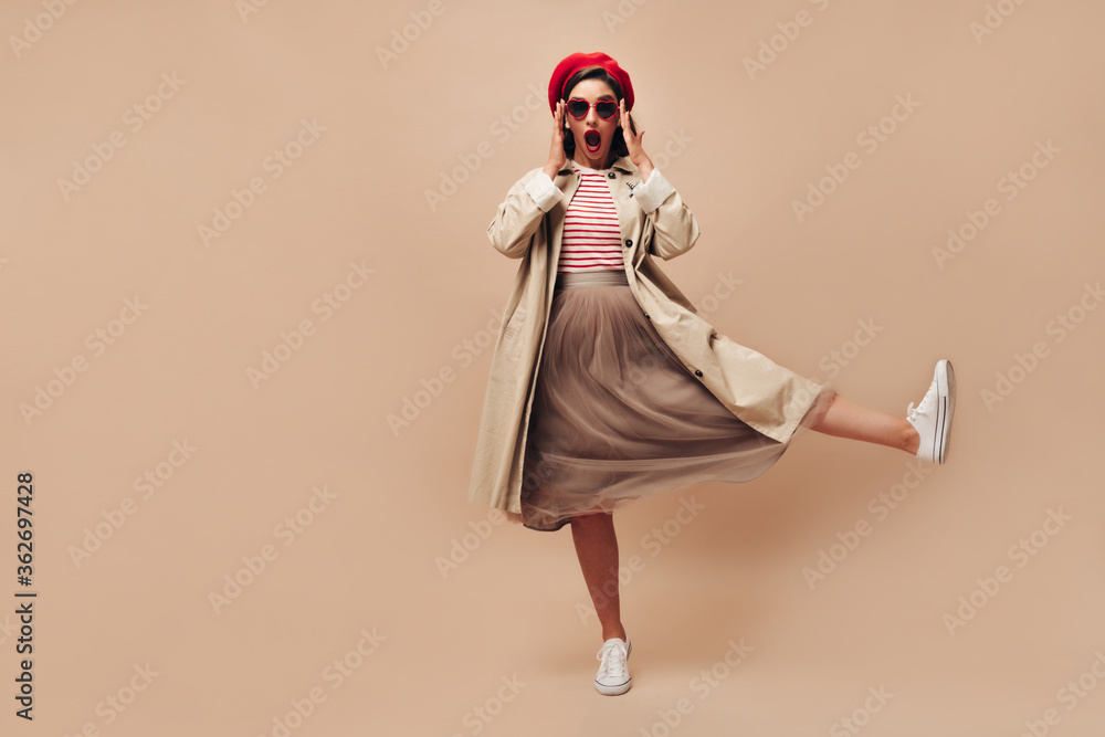 Woman in eyeglasses surprised looks into camera on beige background. Stylish girl in sunglasses in shape of heart and in long skirt raises her leg..