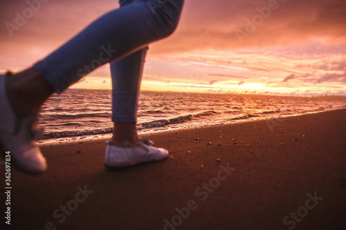 A side view a girl walks alone on the beach at the colorful sunset after the rain. Legs in the white shoes. Orange, crimson purple colours.