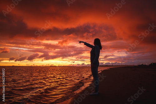Woman walks alone on the beach and looks at the colorful sunset after the rain. Legs in the white shoes. Red orange, crimson purple colours.