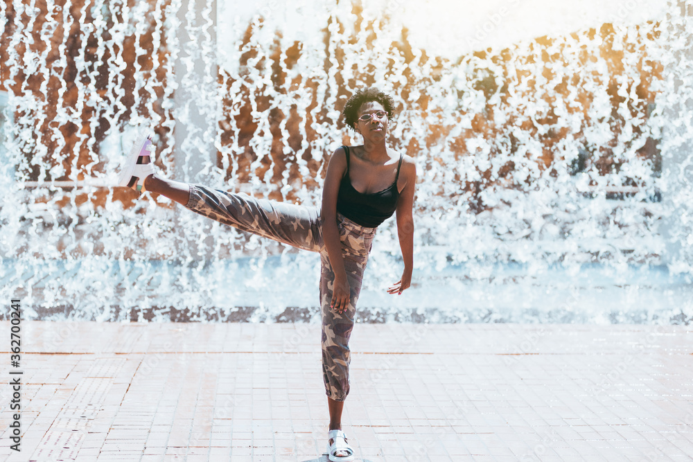 Funny shot with a fancy charming black girl in a sunglasses fooling around, standing on one leg with another leg lifted up doing ballet pas; streams of falling water in background and splashes around