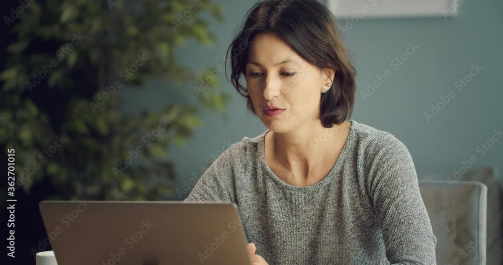 Serious woman dressed in domestic clothing, sitting at table and talking through video chat on wireless laptop. Concept of technology, online communication and social distance. Serious woman dressed in domestic clothing, sitting at table and talking through video chat on wireless laptop. Concept of technology, online communication and social distance.