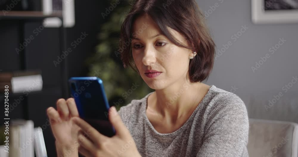 Beautiful woman with dark hair using smartphone while sitting at table. Happy female in domestic outfit spending leisure time with modern gadget. Beautiful woman with dark hair using smartphone while sitting at table. Happy female in domestic outfit spending leisure time with modern gadget.