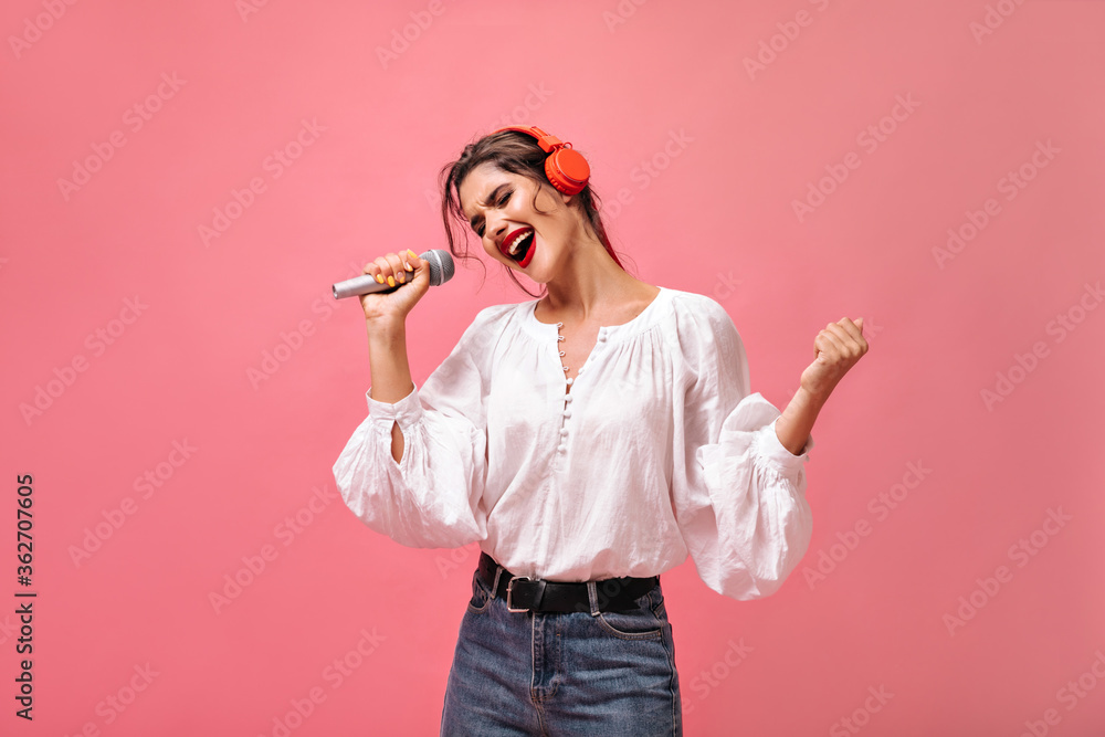 Charming woman in white blouse and headphones singing on pink background. Beautiful woman in stylish dress with red lips posing.. Charming woman in white blouse and headphones singing on pink background. Beautiful woman in stylish dress with red lips posing..