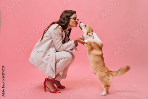 Pretty woman in beige outfit plays with dog on pink background. Cute business lady in stylish suit and red shoes kisses corgi..