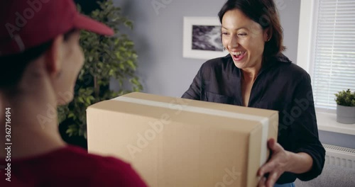 Back view of female courier in red uniform giving cardboard box to beautiful dark-haired woman. Happy client looking very excited while receiving long-awaited order.