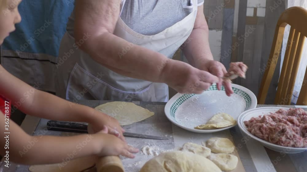 Funny blond boy helps beloved grandmother in kitchen. elderly woman rolls pastry for pasties and puts minced meat. teenager touches dough. Kid and granny are happy together. shallow depth of field Funny blond boy helps beloved grandmother in kitchen. elderly woman rolls pastry for pasties and puts minced meat. teenager touches dough. Kid and granny are happy together. shallow depth of field