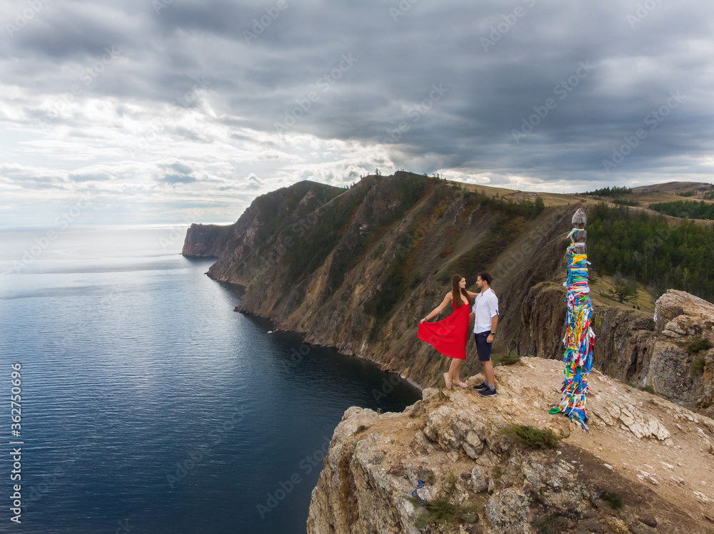 Drone capture of young couple standing on the edge of cliff near spirit pole (Pagan burial) on Olkhon island, Russia. Red dress. Shamanism.