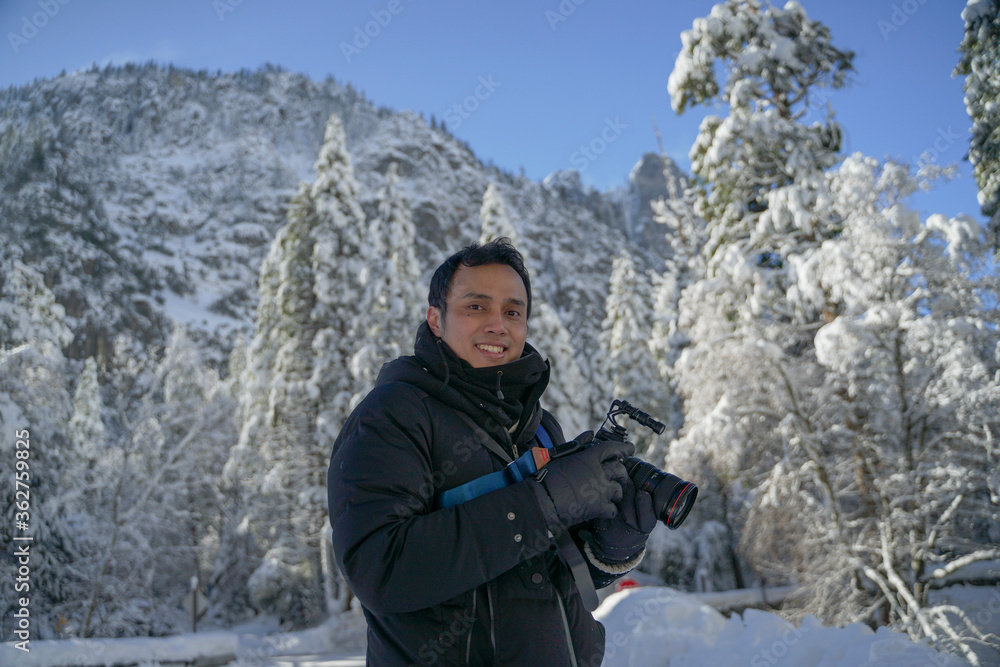 Asian photographer with his professional camera posing smiling with the wonderful Yosemite landscape in winter snow as a background
