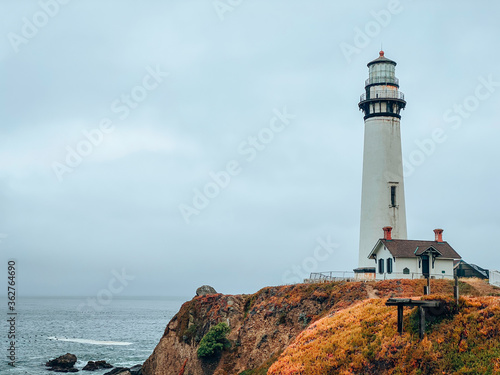 Pigeon point Lighthouse in Pescadero. White lighthouse on the Pacific coast. The tragic dark sky. Colorful flowers. Fall, summer. Free space for text. California, USA