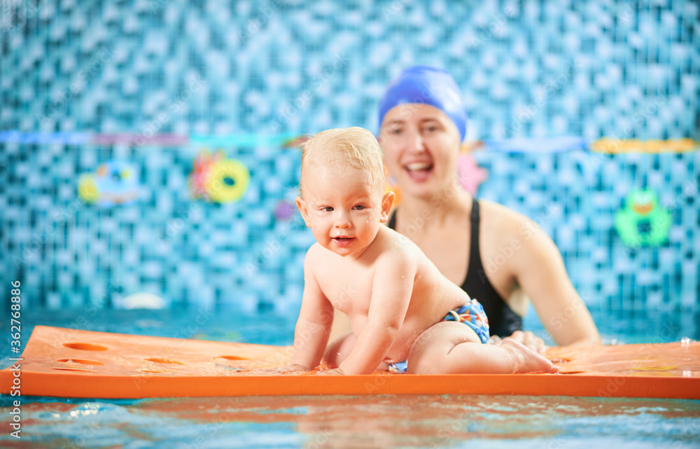 Horizontal snapshot of a happy mother and son spending fun time in swimming pool. A boy is crawling on a floating mat, mother is behind him supporting. Concept of active leisure