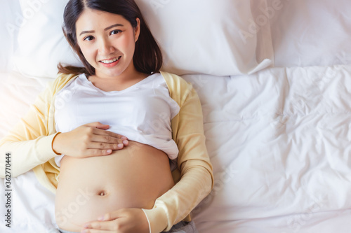 Happy pregnant young asian woman touching belly and admiring her baby or fetus on bed with happiness smile face. Young Asia mother love her baby so much. Mom waiting and expecting baby has good health