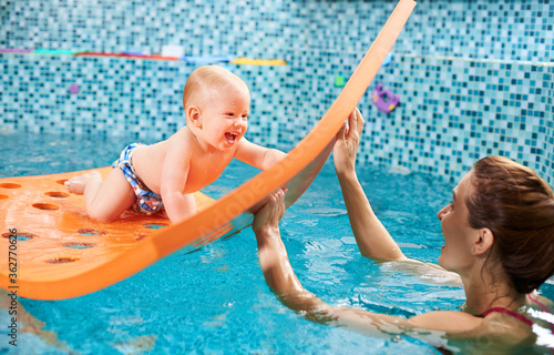 Snapshot of a very happy and playful swimming class with instructor. Cheerful baby boy is crawling on a floating mat with holes and playing with a caregiver