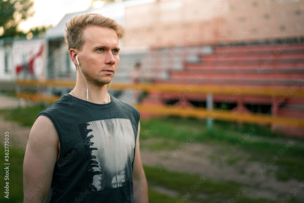 Portrait of a Young man with headphones in his ears who looks at the stadium against the background of empty stands, ready to train and will to win Portrait of a Young man with headphones in his ears who looks at the stadium against the background of empty stands, ready to train and will to win