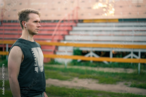 Portrait of a Young man with headphones in his ears who looks at the stadium against the background of empty stands, ready to train and will to win