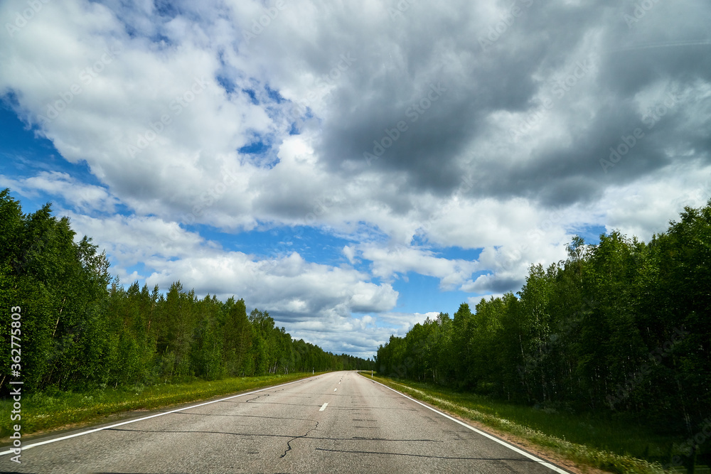 View from relief car windscreen on the blue sky with white clouds, grey asphalt road and landscape with forest and green teeses. Landscape through window