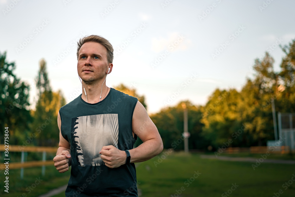 Fototapeta premium Portrait of a young man in fitness wear running in a park. Close up of a smiling man running while listening to music using earphones.