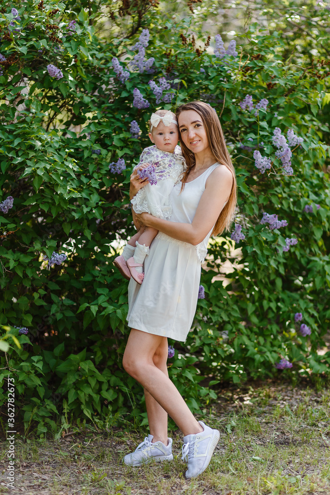 Slender beautiful young mother holds in her arms a baby girl in a white dress in full height. Slender beautiful young mother holds in her arms a baby girl in a white dress in full height.