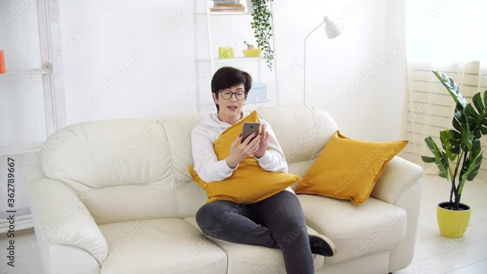 Self isolation concept. Middle-aged woman sitting on a sofa in the living room and talking via video call. Self isolation concept. Middle-aged woman sitting on a sofa in the living room and talking via video call.