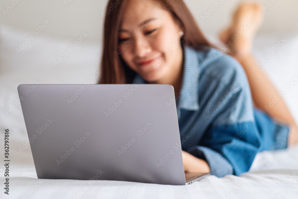 A beautiful asian woman using and working on laptop computer while lying down on a white cozy bed at home A beautiful asian woman using and working on laptop computer while lying down on a white cozy bed at home
