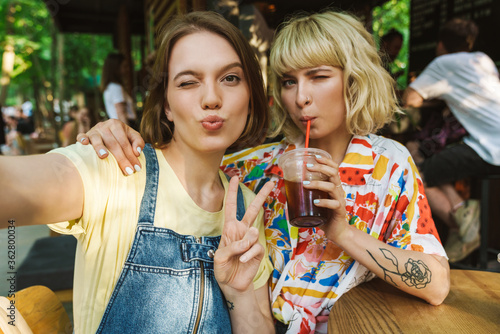 Image of women taking selfie and gesturing peace sign while drinking soda