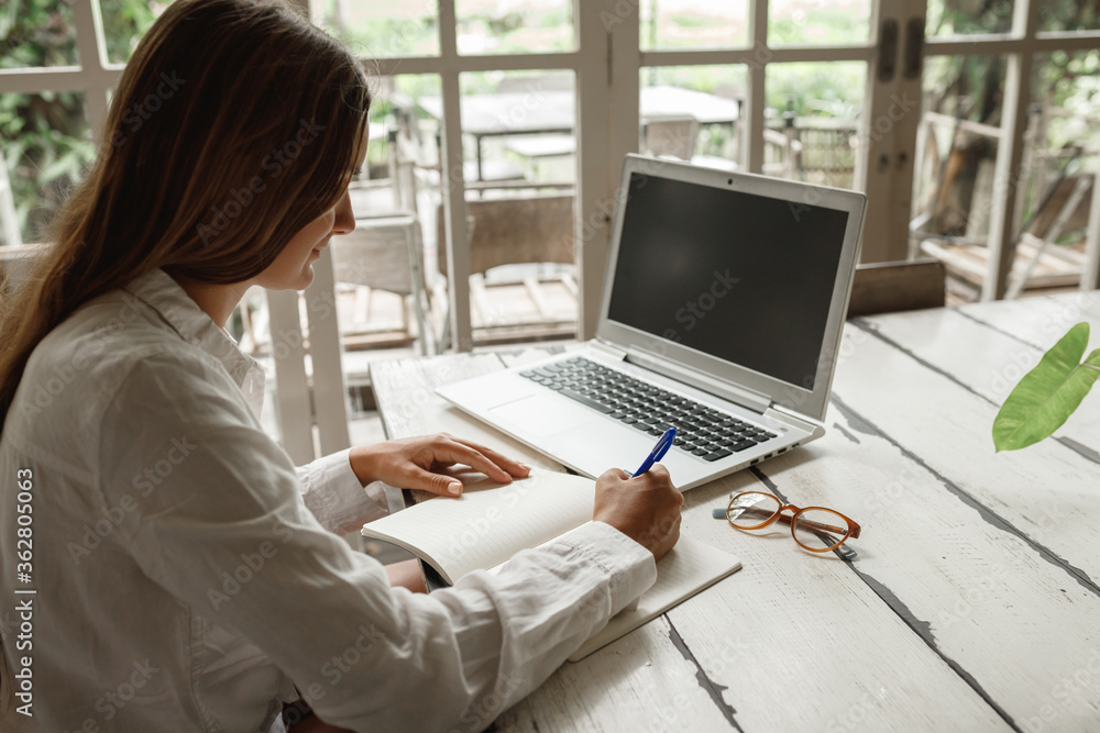 Concentrated at work. Confident young woman in smart casual wear working on laptop while sitting near window in creative office or cafe,writing in the notepad Concentrated at work. Confident young woman in smart casual wear working on laptop while sitting near window in creative office or cafe,writing in the notepad