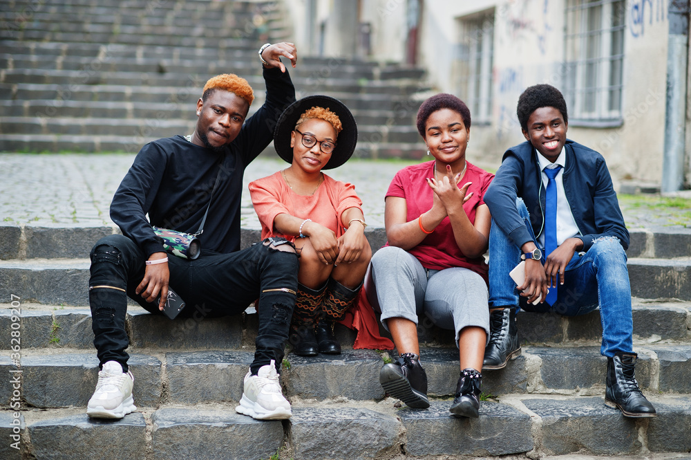 Fototapeta premium Four african friends having fun outdoors. Two black girls with guys sit on stairs of an old city.
