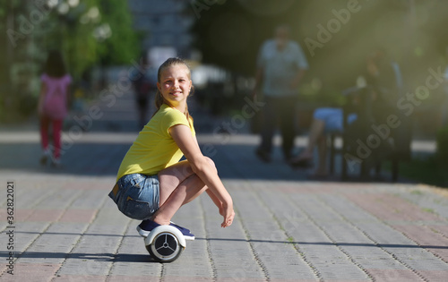 Girl child rides while sitting on a hoverboard in a summer park, happy looking at the camera