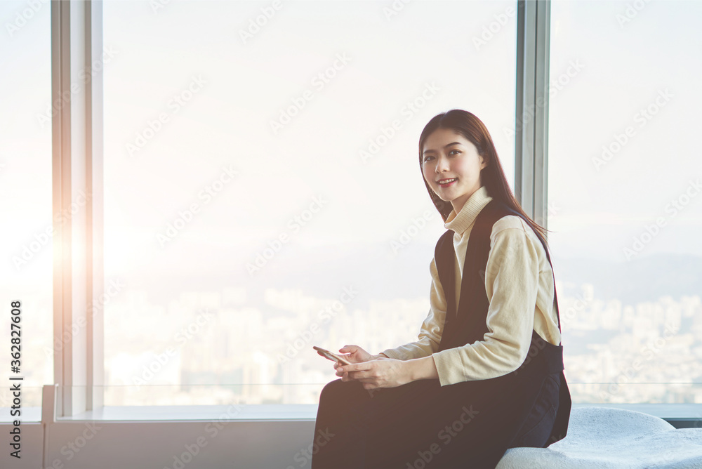 Attractive hipster girl is smiling for someone while is holding smart phone in hands. Chinese female is sitting in modern interior against skyscraper window with copy space background for your content Attractive hipster girl is smiling for someone while is holding smart phone in hands. Chinese female is sitting in modern interior against skyscraper window with copy space background for your content