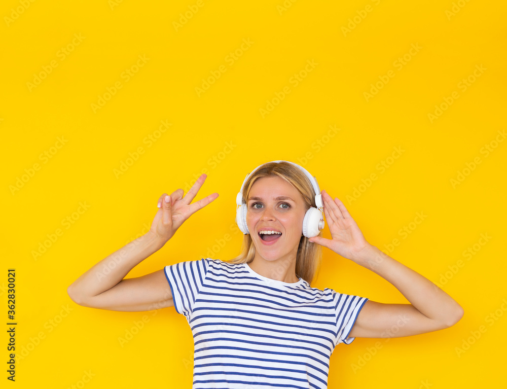 Joyful teen girl with headphones smiling showing peace gesture over yellow background, copy space Joyful teen girl with headphones smiling showing peace gesture over yellow background, copy space
