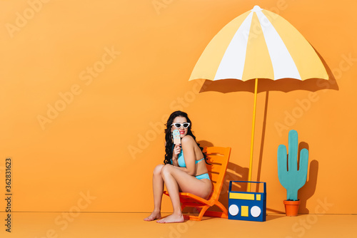 happy woman in sunglasses and swimsuit sitting on deck chair near boombox and umbrella while holding paper ice cream on orange