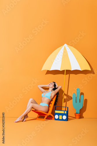 happy woman with closed eyes sitting on deck chair near paper boombox, cactus and umbrella on orange