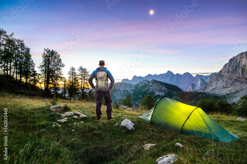 Hiker with Backpack standing on Mountain and Tent glowing under a moon night sky at sunset or sunrise twilight hour. Alps, Sleme, Triglav National Park, Slovenia. photo