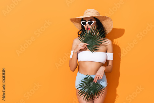 stylish woman in swimsuit, sunglasses and straw hat holding palm leaves on orange