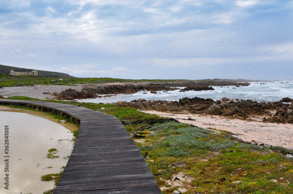 Wooden boardwalk near the rocky coast and blue ocean with waves on the windy day at Cape Agulhas, the most southern point of Africa, where the Indian and Atlantic oceans meet, South Africa