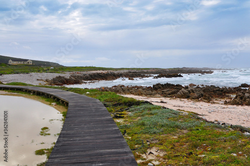 Wooden boardwalk near the rocky coast and blue ocean with waves on the windy day at Cape Agulhas  the most southern point of Africa  where the Indian and Atlantic oceans meet  South Africa