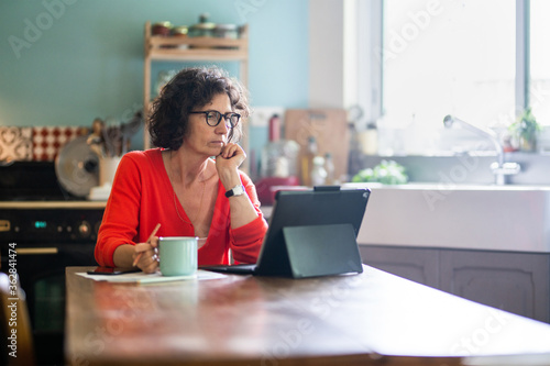 Billede på lærred Beautiful middle-aged woman sitting in her kitchen, working on her laptop while drinking tea